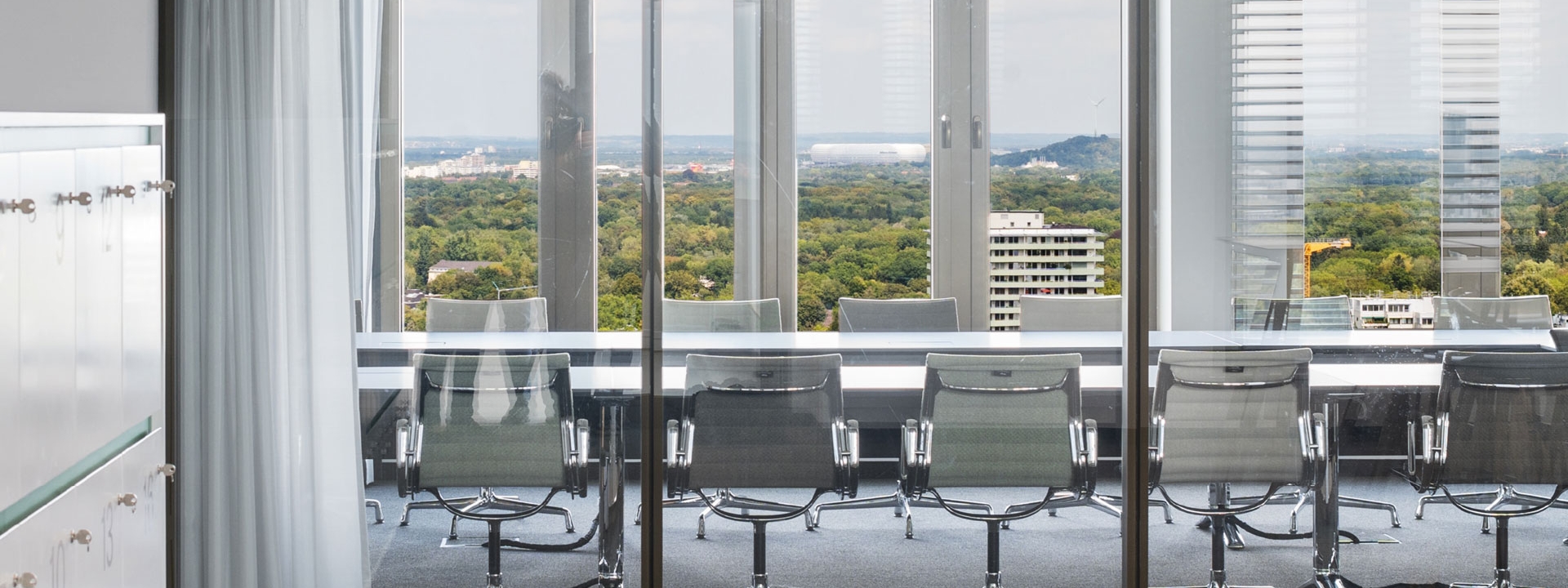 Blick durch die Glasscheibe in den Tagungsraum und im Hintergrund die Aussicht auf Wald und die Allianz Arena in München